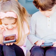 Två barn sitter med böjda huvuden över varsin mobil. En flicka med rosa rosett och en pojke i grå tröja. De verkar drabbade av "tech neck".