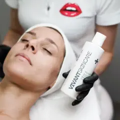En kvinna med sluten blick får en ansiktsbehandling. En hand med svart handske håller upp en flaska VIVANT SKINCARE, kanske för att behandla fet hy.