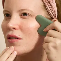 Kvinna med rosa pannband använder en grön gua sha för ansiktet, med fingrarna som stödjer hakan.