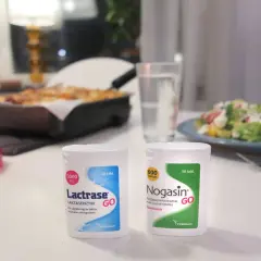 Två förpackningar laktos tabletter, Lactrase och Nogasin, står på ett bord med mat i bakgrunden.