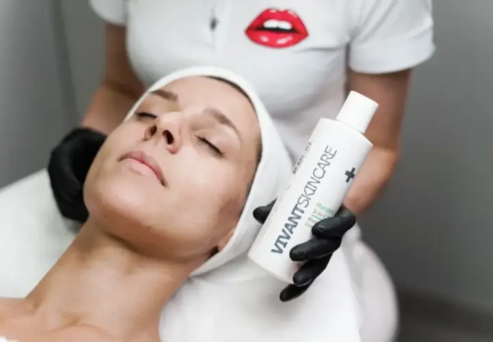 En kvinna med sluten blick får en ansiktsbehandling. En hand med svart handske håller upp en flaska VIVANT SKINCARE, kanske för att behandla fet hy.