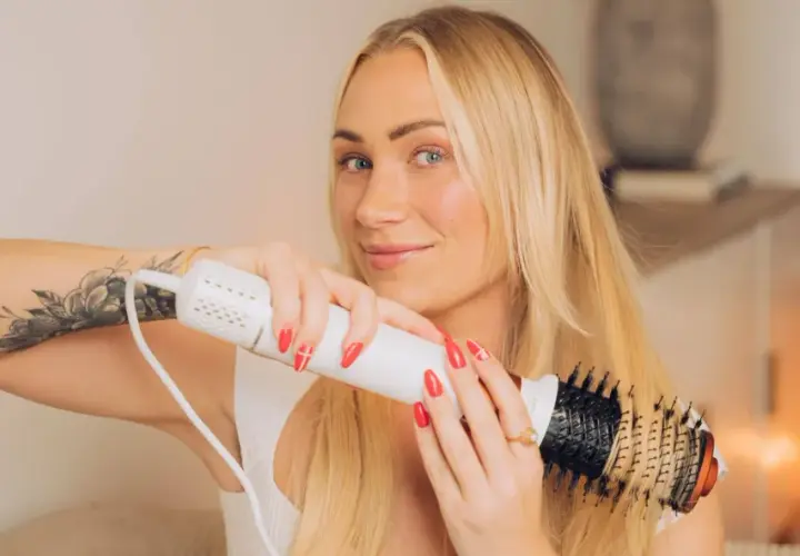 Blond kvinna med tatuering på armen stylar sitt hår med en varmluftsborste för perfekt blow out hår.