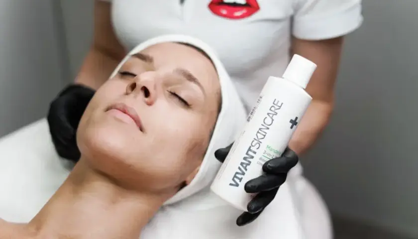 En kvinna med sluten blick får en ansiktsbehandling. En hand med svart handske håller upp en flaska VIVANT SKINCARE, kanske för att behandla fet hy.