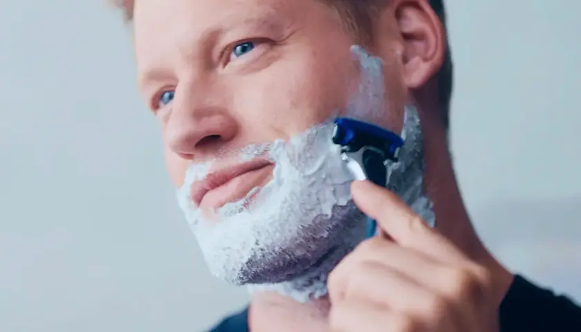 Man med blå ögon raka med kniv och raklödder på kinden.