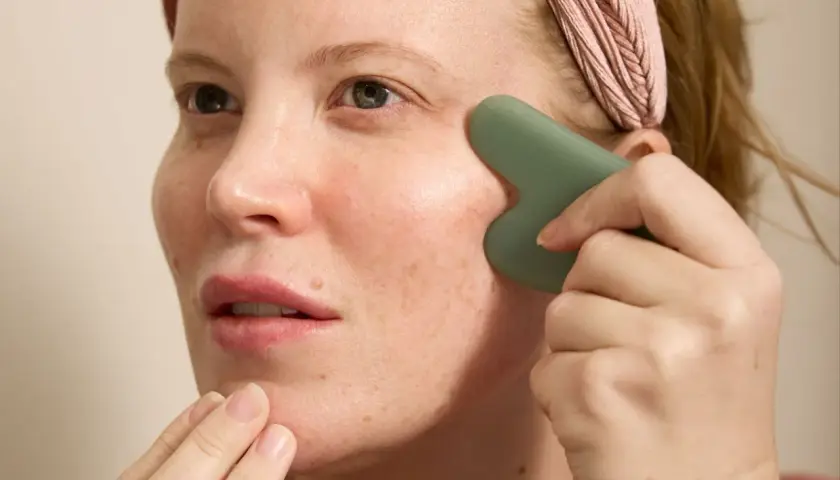 Kvinna med rosa pannband använder en grön gua sha för ansiktet, med fingrarna som stödjer hakan.