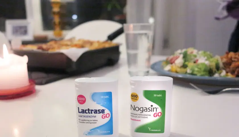 Två förpackningar laktos tabletter, Lactrase och Nogasin, står på ett bord med mat i bakgrunden.