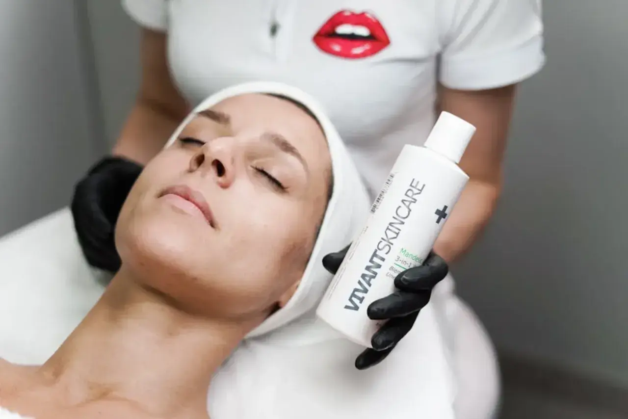 En kvinna med sluten blick får en ansiktsbehandling. En hand med svart handske håller upp en flaska VIVANT SKINCARE, kanske för att behandla fet hy.