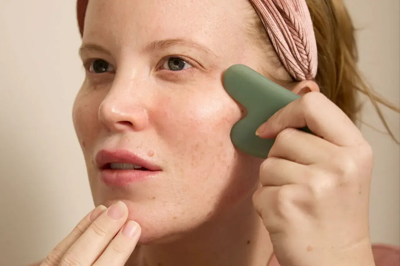 Kvinna med rosa pannband använder en grön gua sha för ansiktet, med fingrarna som stödjer hakan.