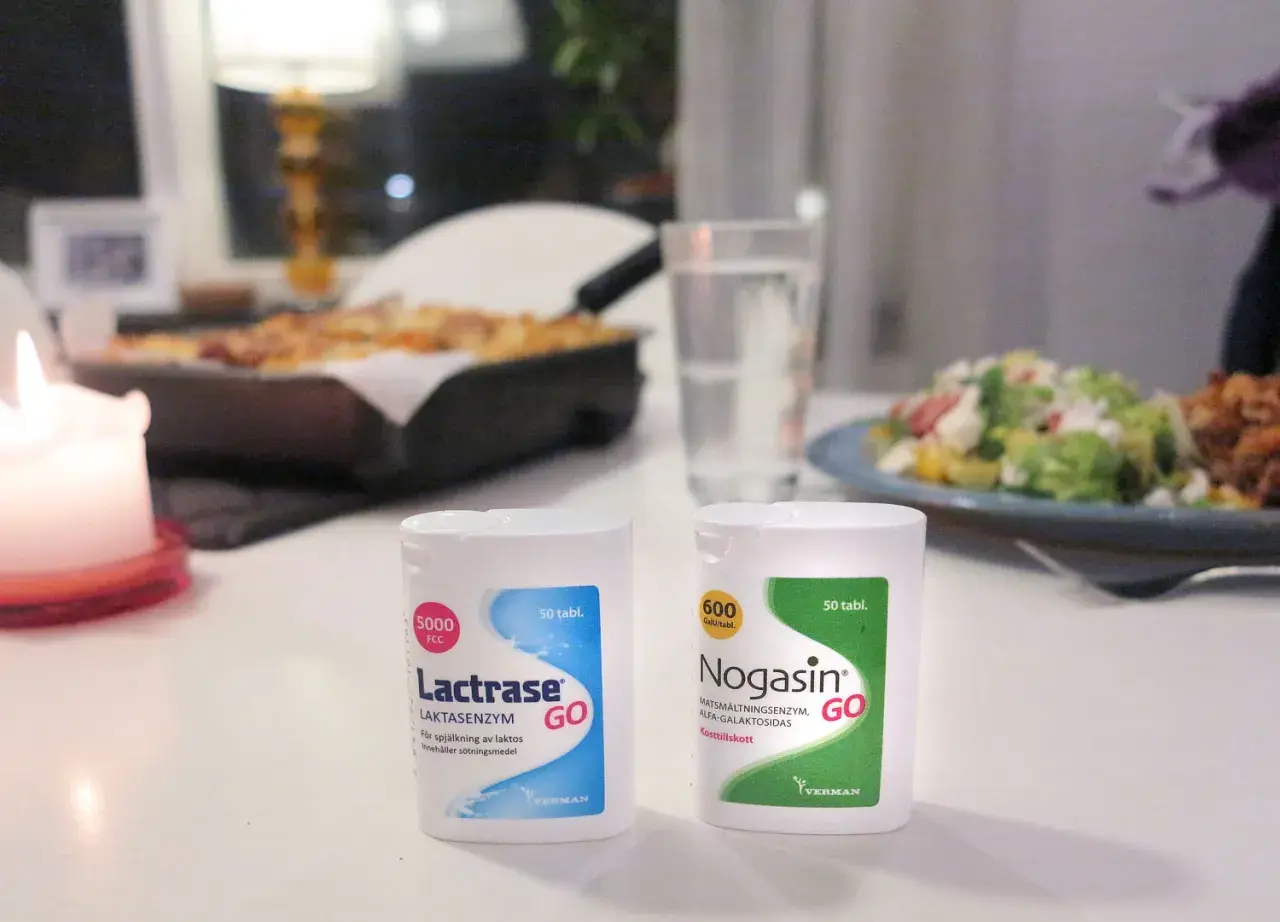 Två förpackningar laktos tabletter, Lactrase och Nogasin, står på ett bord med mat i bakgrunden.