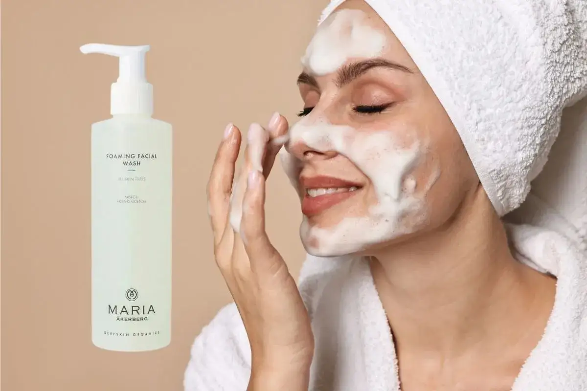 En kvinna med skum i ansiktet anv&auml;nder Maria &Aring;kerberg foaming facial wash. En flaska av produkten syns till v&auml;nster.