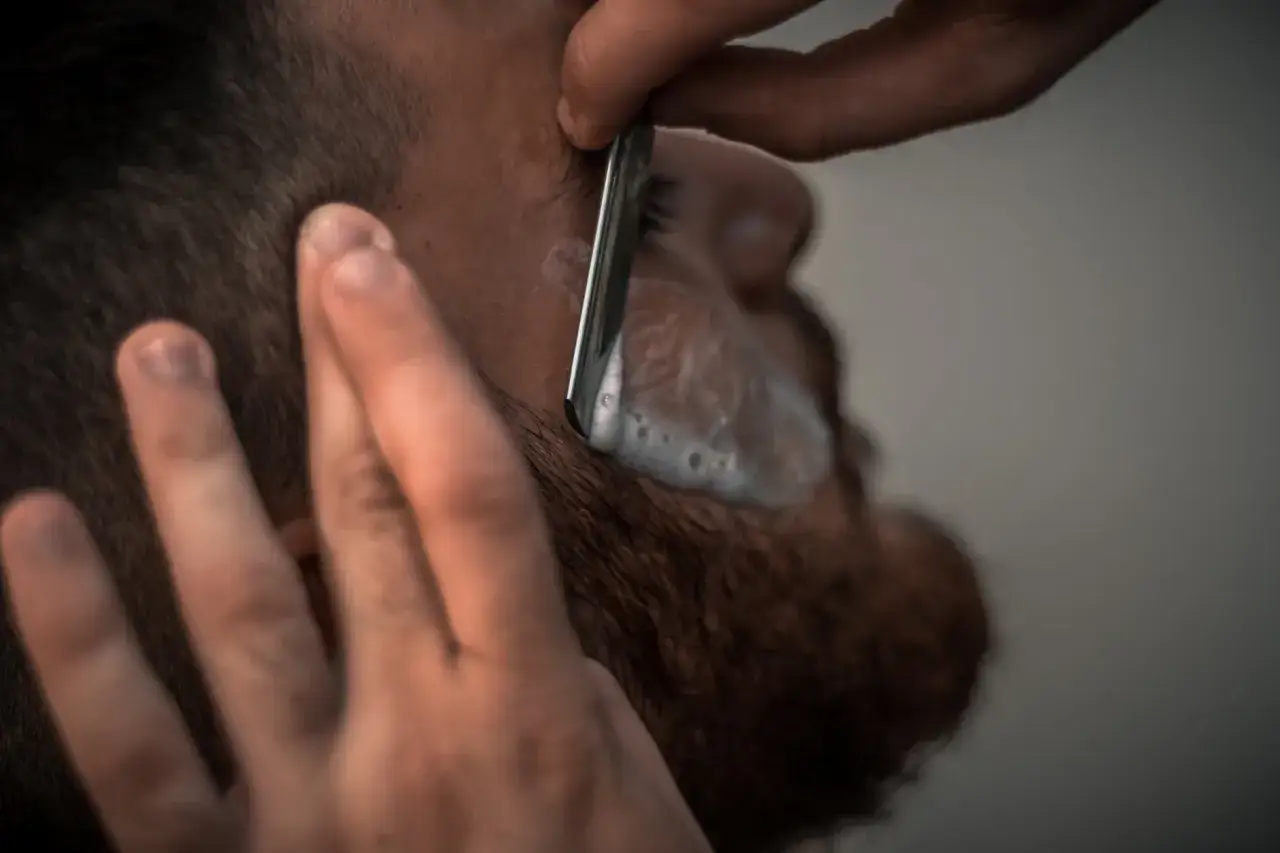 En barberare använder en rakkniv för att raka med kniv. Skummet och skäggstubben syns tydligt.